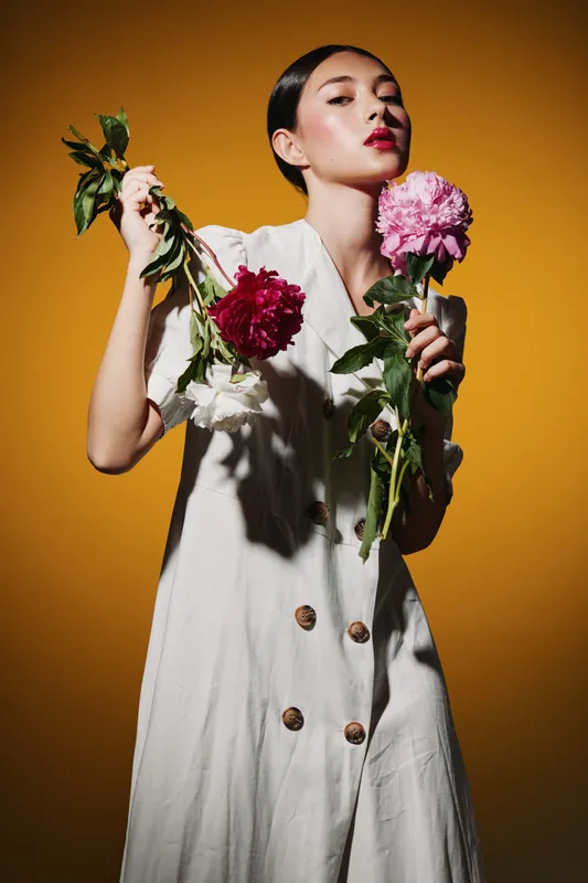 Elegant woman with bold red lipstick and sleek hair conveys beauty and fashion creativity while holding vibrant colorful flowers against a warm orange background.