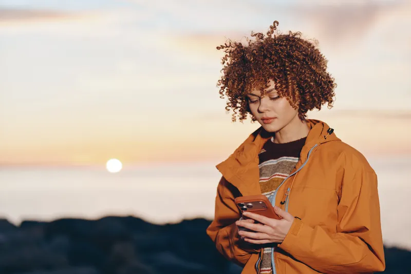 Woman with curly hair standing outdoors at sunset, wearing orange jacket, holding a tablet, casual lifestyle moment, warm golden light, soft wind, calm confident expression