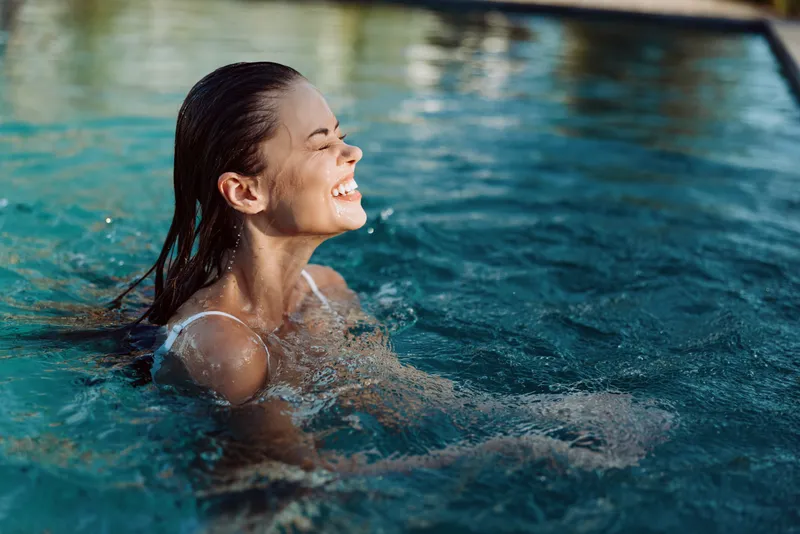 Joyful woman in a pool, smiling with eyes closed, sunlit water and gentle ripples, relaxed posture, bright blues, carefree mood, natural lifestyle moment