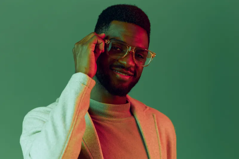 Stylish young man with glasses smiling and posing against a green background, showcasing contemporary fashion and confidence