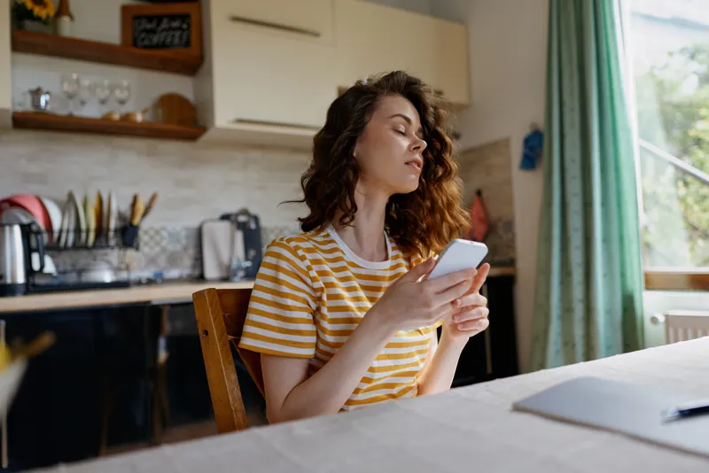 Smiling woman in a striped yellow shirt using a smartphone at a kitchen table, indoors with daylight streaming through a window, capturing a casual home lifestyle moment.