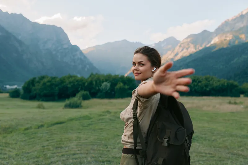 Young woman with a backpack extends her arm toward the camera in a wide valley with towering mountains, inviting a sense of freedom and adventure in open field today.