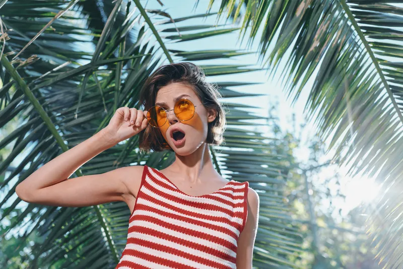 A cheerful woman wearing a red and white striped sleeveless top and orange sunglasses stands beneath palm fronds, sunlit and relaxed, creating a vibrant lifestyle moment in a tropical setting.