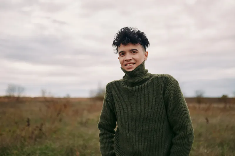 Authentic portrait of a young man outdoors in a thick knit sweater, presenting a candid, credible moment. A calm field, soft light, and subtle smile convey sincerity.