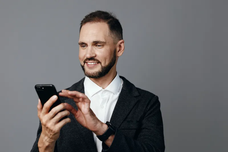 A professional man in business attire uses a smartphone against a solid colored background, exuding calm confidence, focused determination, and deliberate presence.