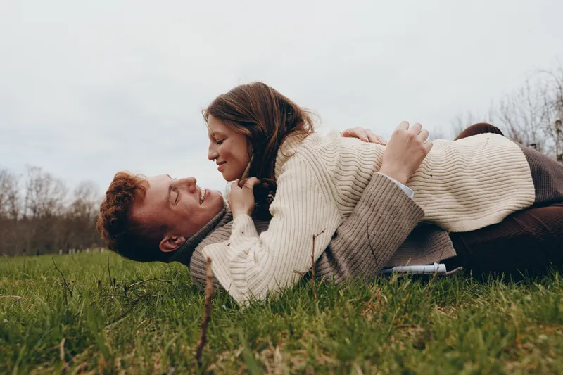 A young couple in cozy sweaters lies on the grass outdoors sharing a tender moment and smiling warmly at each other during a relaxed lifestyle scene.