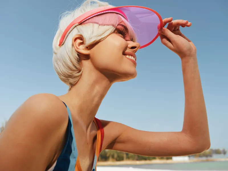 A smiling woman on a sunny beach wears a pink visor and sporty swimwear, posing in profile with a light breeze and clear blue sky creating a vibrant summer scene.