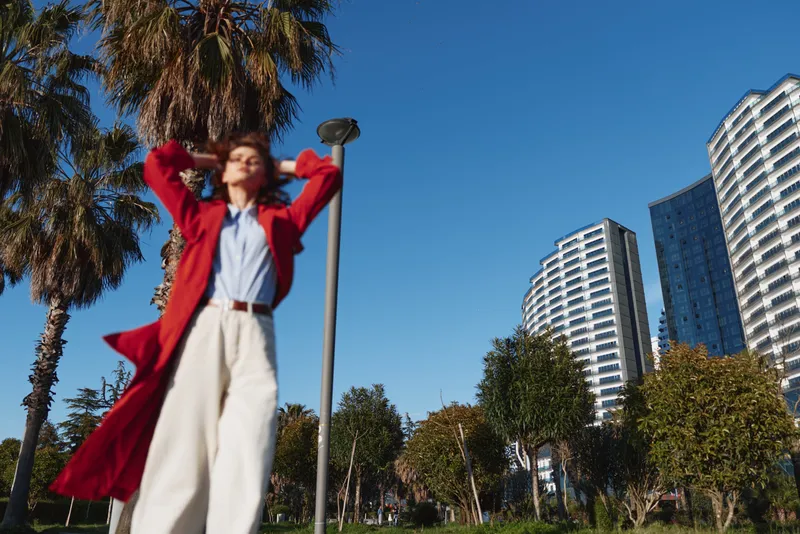 A confident woman in a red coat stands outdoors with her hands behind her head, surrounded by palm trees and modern buildings under a clear blue sky.