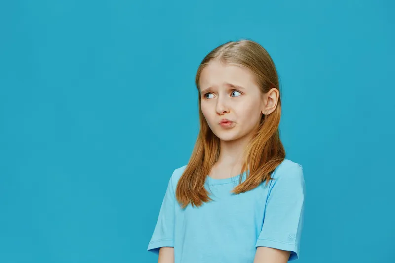 A young girl with shoulder length blonde hair, wearing a light blue t shirt, displays a thoughtful expression against a bright blue background, embodying curiosity and innocence