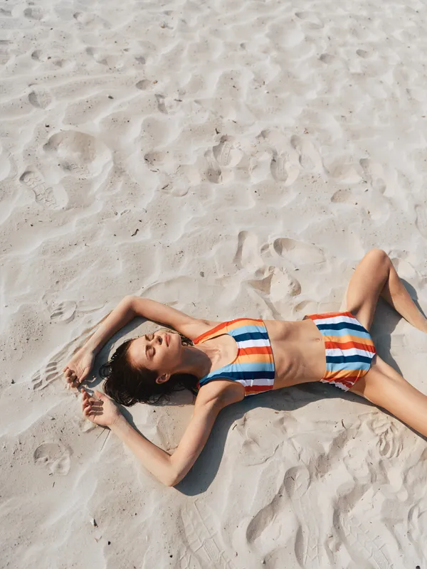 Young woman in a striped bikini reclines on sunlit sand, arms raised over head, enjoying a calm beach moment under bright daylight and a warm sea breeze.