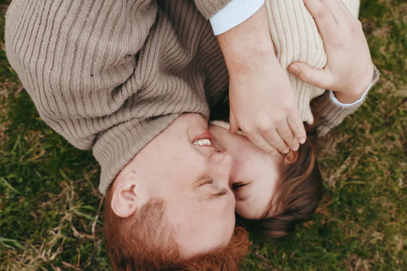 Joyful couple outdoors, hugging on grass, sunlit moment, warm knit sweaters, intimate connection, natural colors, breeze, relaxed pose, candid embrace, carefree happiness