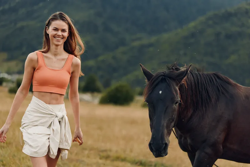 A joyful young woman walks beside a dark horse in a sunlit meadow, smiling warmly as she enjoys a peaceful countryside moment with gentle weather, companionship, and natural harmony.