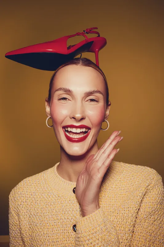 Smiling woman with red high heel shoe balanced on her head wears yellow sweater and gold hoop earrings. Studio portrait on brown background with playful and joyful mood concept.
