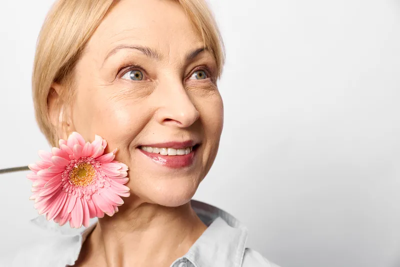 Woman elderly smiling portrait with pink flower near cheek, soft studio light, gentle expression, beauty concept for skincare and anti aging, spring mood, copy space on white background