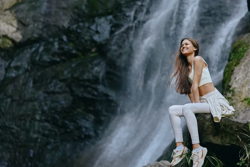 Smiling woman sitting near a large waterfall in casual white clothing, enjoying nature and peaceful outdoor adventure with a joyful expression.