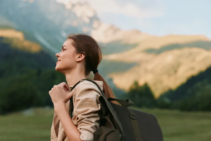 Woman hiker with backpack stands outdoors, gazing at distant mountains, enjoying nature and fresh air, serene lifestyle moment in warm sunlight under a clear sky and peaceful mood of exploration