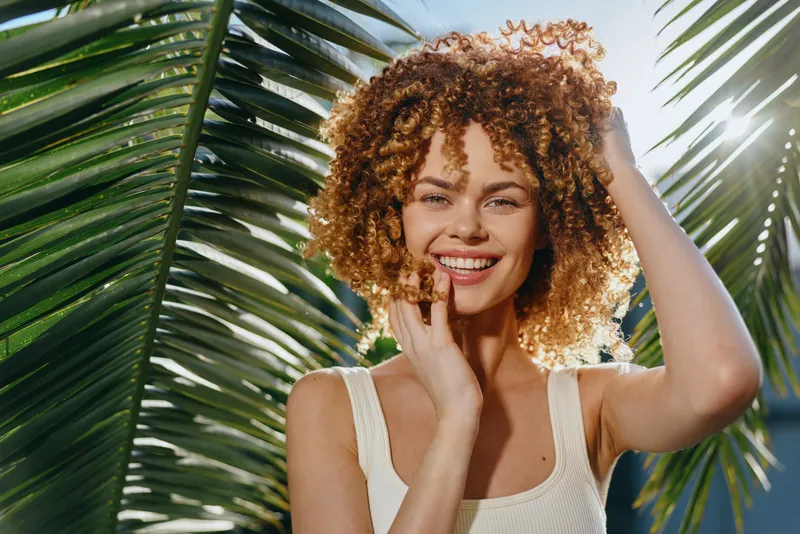 Smiling woman with curly hair enjoys sunny tropical outdoors framed by green palm leaves, wearing light sleeveless top and radiating natural positive energy.