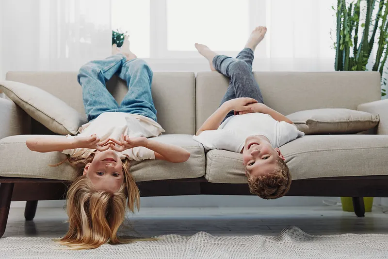 Two happy children lying upside down on a beige couch in a bright living room, smiling and playing together. Concept of childhood, fun, and home comfort.