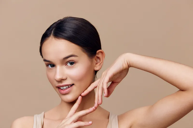 Young female model with a natural smile posing against a beige background, showcasing her skin care routine and healthy lifestyle, embodying beauty and confidence