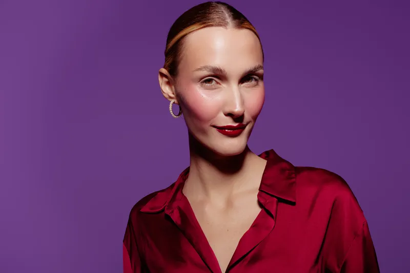 Confident woman with slicked back hair wears red satin blouse and hoop earrings against purple background. Studio portrait with soft lighting highlights facial features and elegant style.