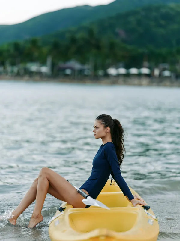 A young woman sits on a yellow kayak in calm coastal water, wearing a dark blue wetsuit, gazing toward the horizon as distant mountains rise behind the shoreline.