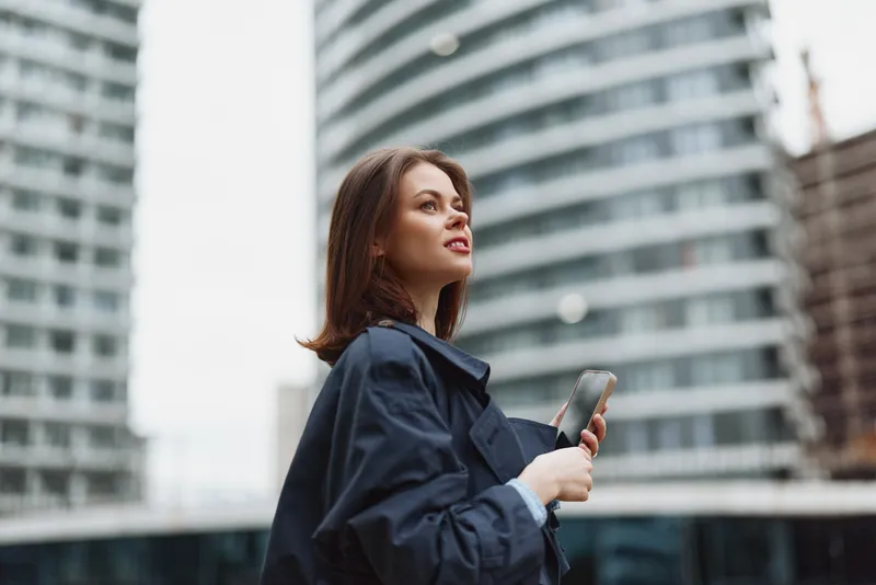 A focused woman in a navy trench coat holds a smartphone, gazing upward in a modern cityscape, conveying ambition and curiosity.