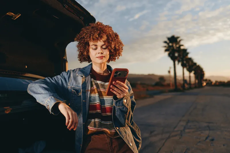 Woman lifestyle outdoors near car, curly hair smiling, holding smartphone at sunset, relaxed pose by open door, warm light, evening breeze, soft clouds, palm trees, late day