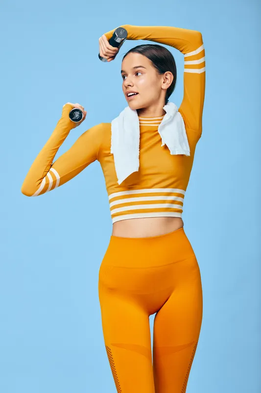 Fitness focused woman in bright orange athletic set lifts dumbbells with a towel around her neck against a blue backdrop, exuding energy, strength and an active lifestyle in a bold workout moment.