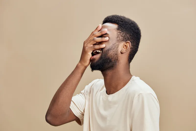 Joyful young man laughing with hand over his face, showcasing happiness against a soft beige background, ideal for emotional expressions in arts