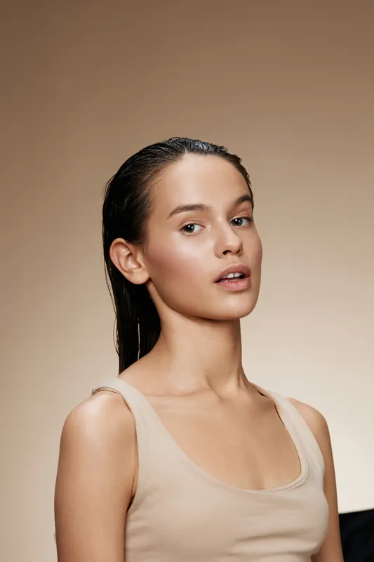 Young woman with wet hair and natural makeup against a warm beige background, showcasing healthy skin and a relaxed expression, ideal for beauty and skincare topics