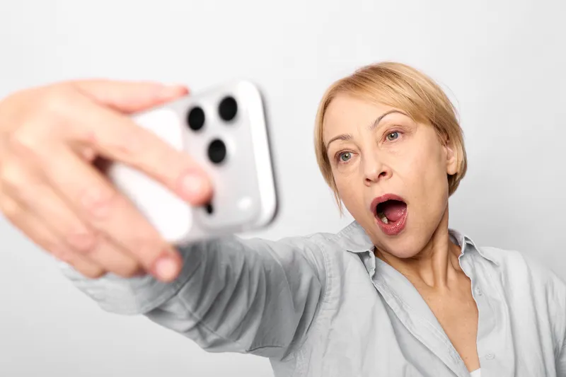 Woman surprised smartphone selfie hand portrait shock mouth open expression, blonde female in gray shirt, indoor white background, close up