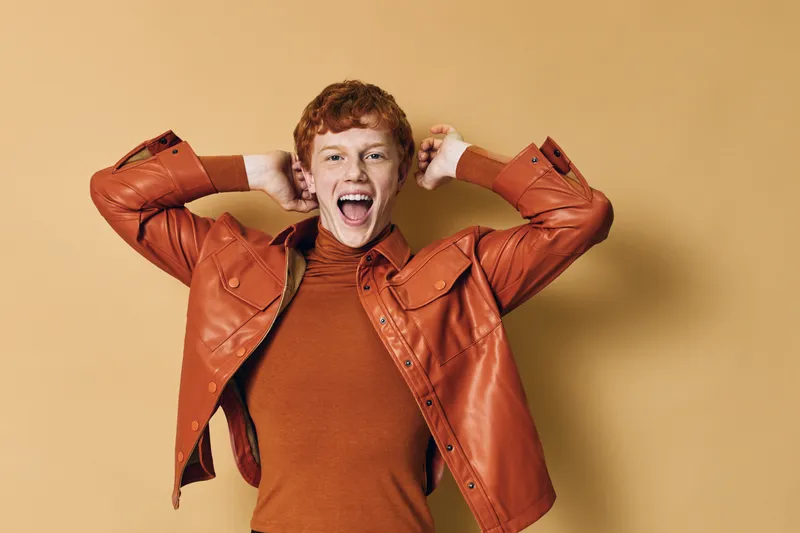 Young redhead man with curly hair wearing orange jacket and sweater posing excitedly with open mouth and hands behind head on beige background.