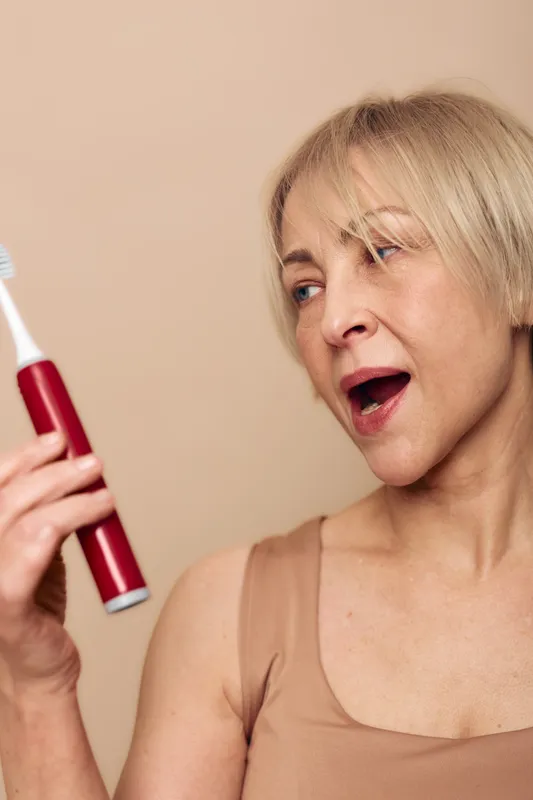 Middle aged woman with short blonde hair holds red electric toothbrush looking at it with open mouth. Concept for oral hygiene, dental care, personal routine, and healthy lifestyle.