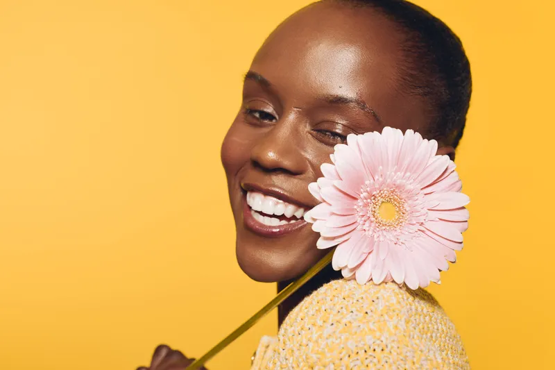 Smiling black woman with dark skin holding pink gerbera flower near face on yellow background. Concept of beauty, happiness, natural skin, and floral wellness in studio portrait.