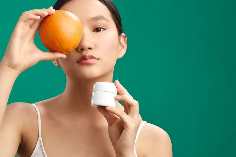 asian woman with a bright expression holds an orange near her eye and a white cream jar, captured in a clean studio setting, conveying freshness, skincare care, and a vibrant beauty routine.