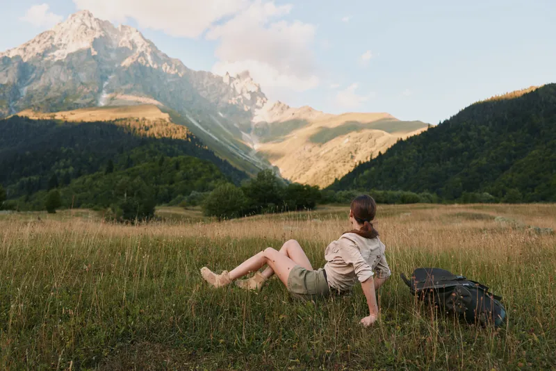 Young man relaxing in a meadow with a backpack as distant mountains rise, bathed in natural light for a serene outdoor moment