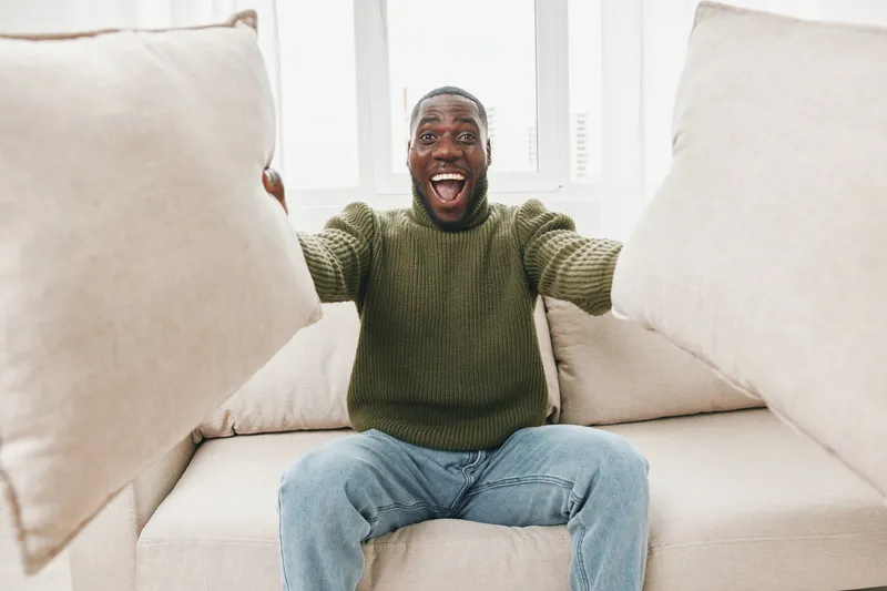 Happy man celebrating at home with pillows, wearing a cozy green sweater and jeans, showcasing joy in a bright living room setting