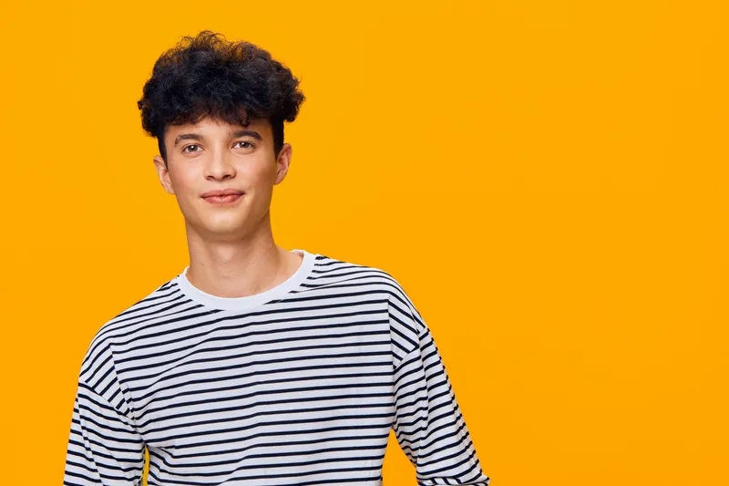 young man with curly hair wearing striped shirt stands against vibrant orange background with relaxed expression and casual pose, showcasing modern youthful style.