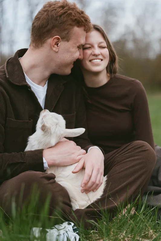 Young couple sitting on grass with a white baby goat in their arms enjoying a tender moment outdoors in casual clothing during a calm spring day.
