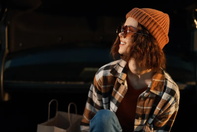 Woman in beanie and sunglasses smiles in a plaid shirt while sitting in a car trunk, candid lifestyle portrait with golden hour glow, authenticity, mindful living and emotional storytelling.