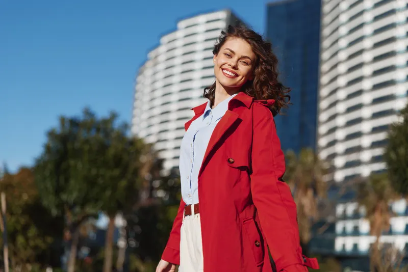 Young woman wearing a red coat outdoors in a sunny urban setting, smiling confidently against modern buildings, urban style, contemporary fashion, and a vibrant city scene.