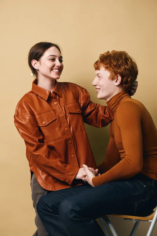 Young couple sitting and holding hands with smiles, woman touching man’s head in affectionate moment with casual clothing, warm tones and indoor studio setting.