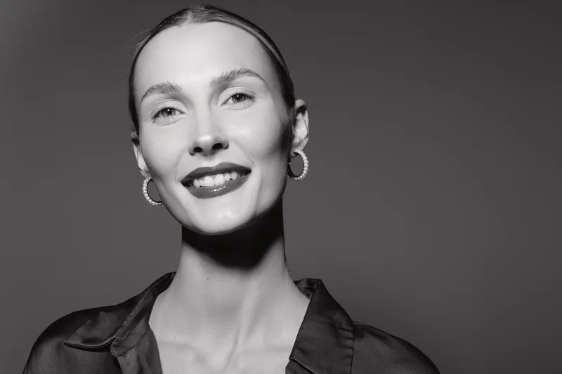 Smiling white woman with light skin and slicked back hair wears hoop earrings and dark blouse, portrait on monochrome background. Concept of beauty and elegance.