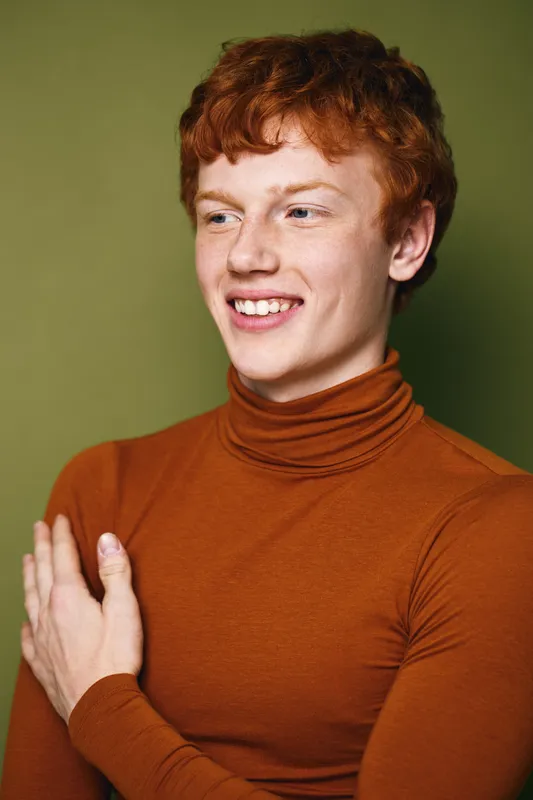 Young man with red hair wearing orange turtleneck sweater smiling with crossed arms on green background in casual stylish outfit indoors.