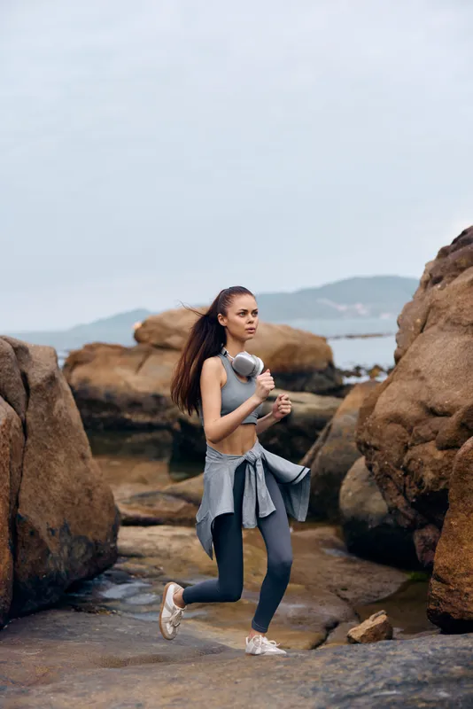 Young athletic woman jogging outdoors near rocky beach, wearing gray sportswear, expressing determination and focus, ideal for fitness and healthy lifestyle concepts