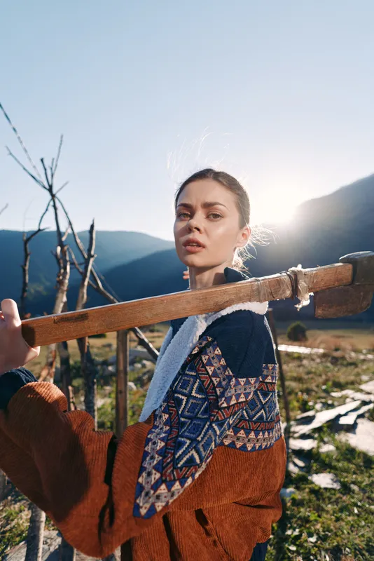 Woman portrait outdoors in mountains wearing patterned sweater, carrying a wooden beam on shoulder with backlit sunlight. Rural landscape, cozy autumn style and confident gaze.