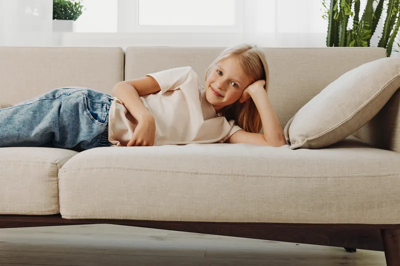 Smiling girl with long blonde hair lies relaxed on beige sofa in bright living room. Cozy home interior with cushions and natural light creates peaceful, comfortable atmosphere for rest.