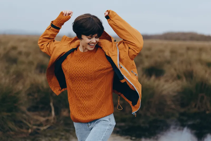 Woman lifestyle outdoors, autumn field scene, orange sweater portrait, smiling with hands raised, adjusting hair, casual jacket, denim jeans, windy countryside, near river, natural daylight
