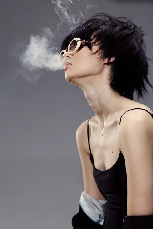 Stylish woman wearing sunglasses exhales smoke with dramatic lighting and minimalist background