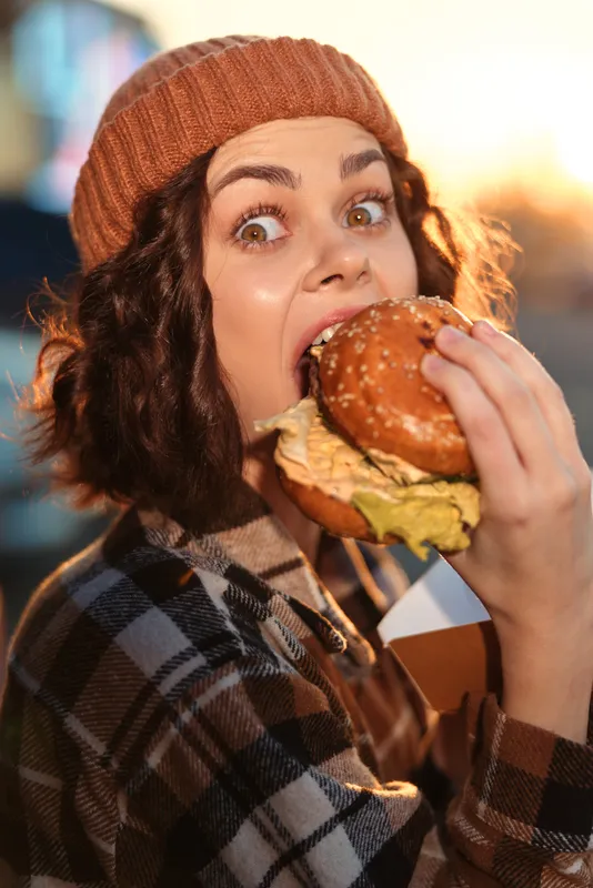 Woman biting a large burger outdoors, beanie and plaid coat in candid moment; authenticity and candid lifestyle captured with golden hour glow, mindful living and emotional storytelling.